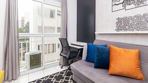 A modern living room with a grey sofa, two colorful cushions, a black office chair, and a large window with a view of buildings. - Vossa Bossa Pinheiros (São Paulo)