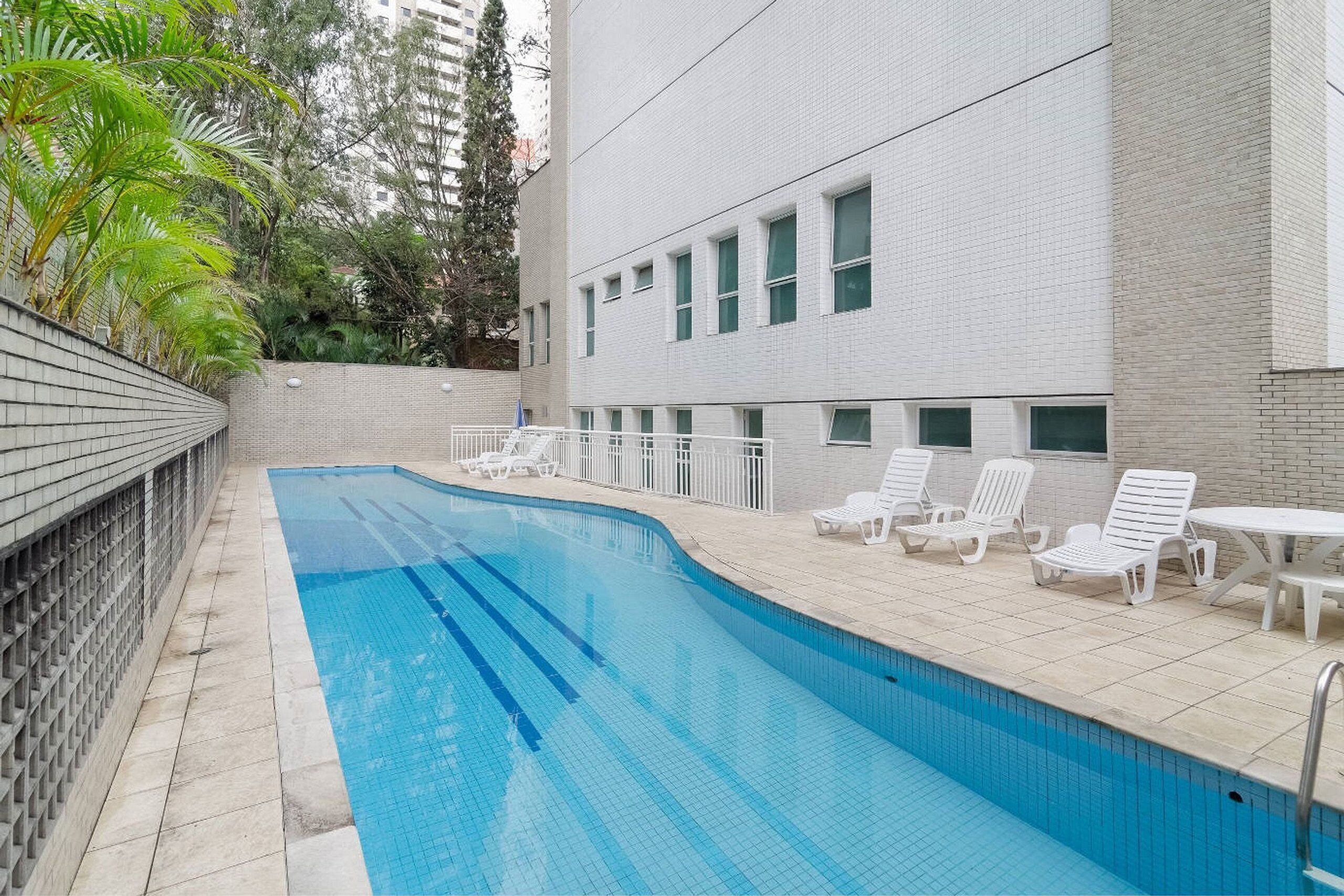 A swimming pool with white lounge chairs and a tiled deck, surrounded by a brick wall and greenery.