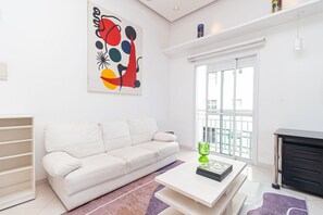 Uma sala de estar moderna com um sofá branco, uma mesa de centro, uma estante de livros e um quadro abstrato colorido na parede.