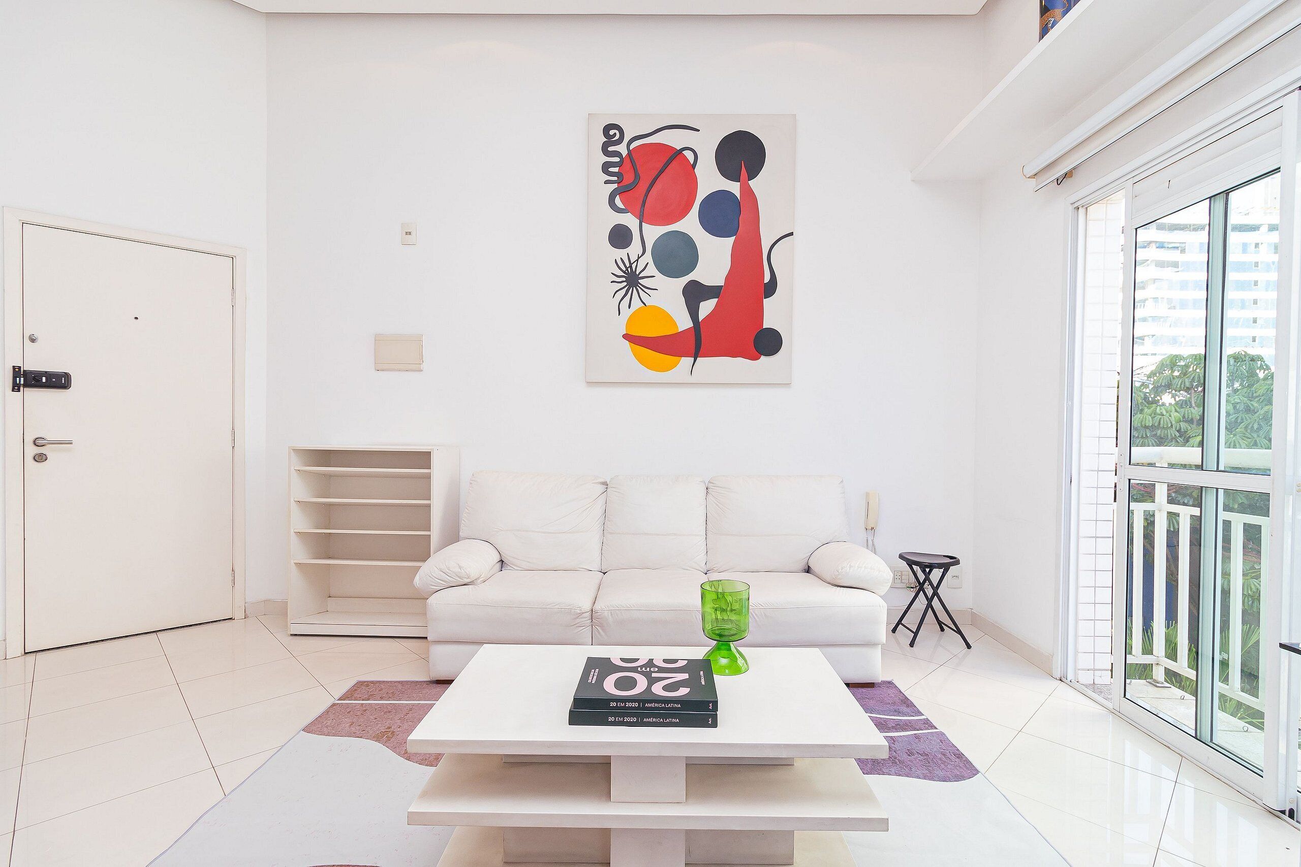 A modern living room with a white sofa, a coffee table with books and a green vase, and a colorful abstract painting on the wall.
