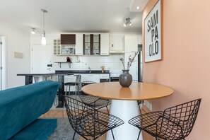 Uma cozinha moderna com armários brancos, uma área de jantar com mesa redonda e cadeiras de arame, e um sofá verde-água.