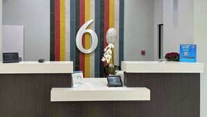 A reception desk with a large number '6' on the wall, a digital screen, a small plant, and a blue sign with a QR code. - Studio 6 Nacogdoches, Tx – Sfa University (Nacogdoches)