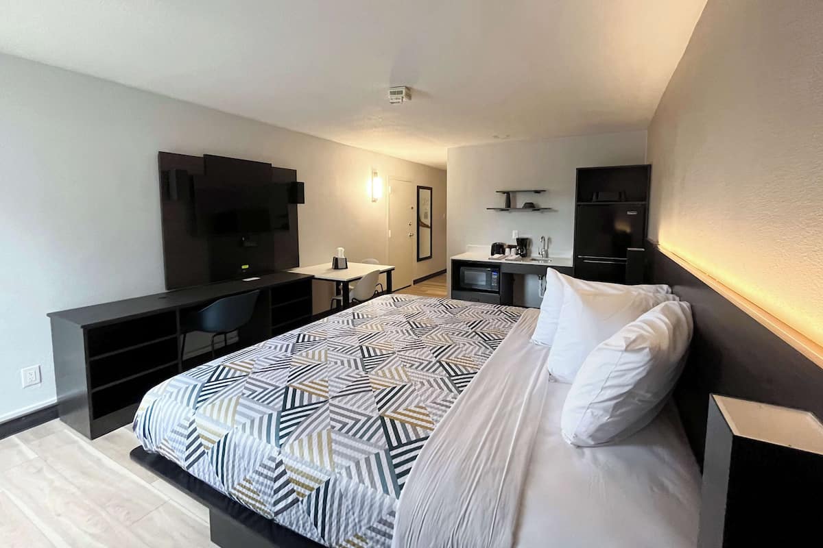 A modern hotel room with a large bed, a desk with a chair, a TV, a microwave, and a refrigerator.