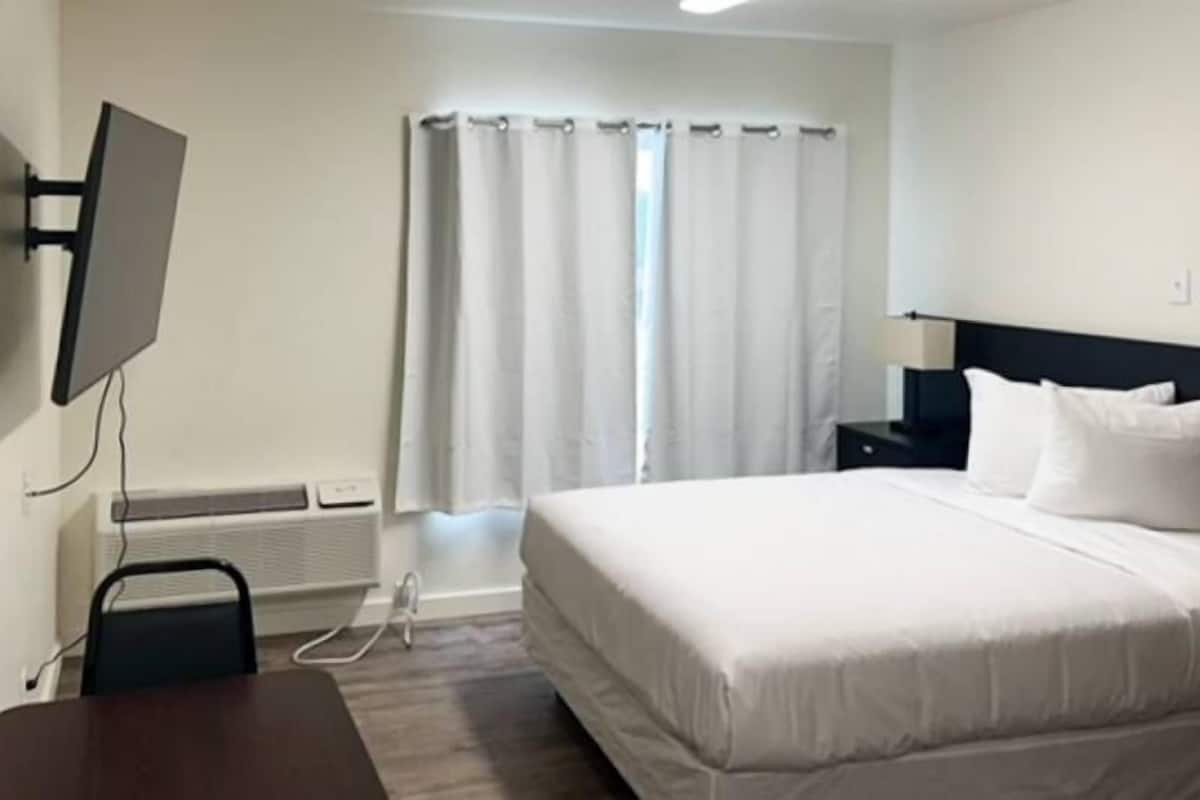 Una habitación de hotel con cama, televisor montado en la pared, mesita de noche y ventana con cortinas.