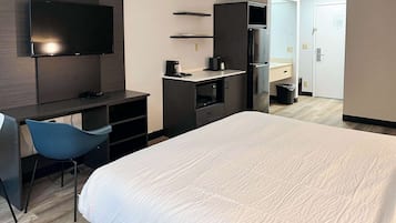 A hotel room with a bed, a desk, a chair, a TV, a microwave, a refrigerator, and a trash bin.