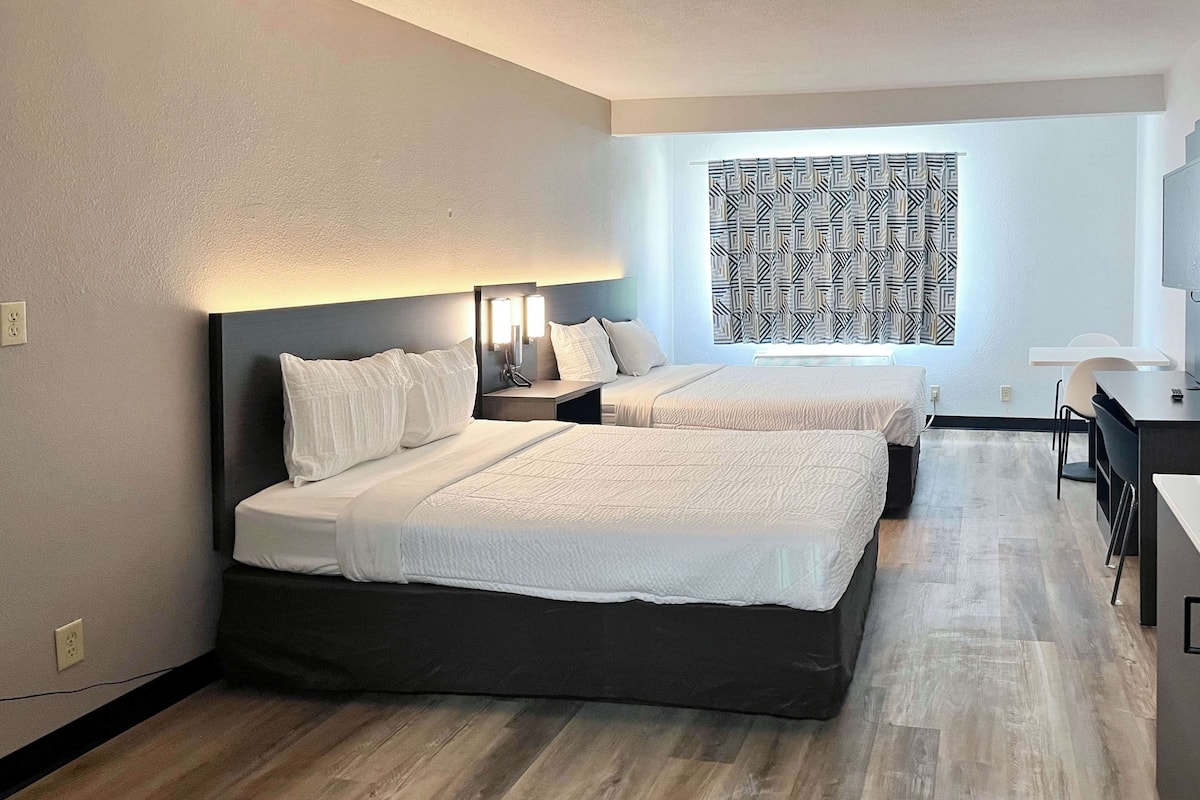 A hotel room with two beds, a desk, and a patterned window.