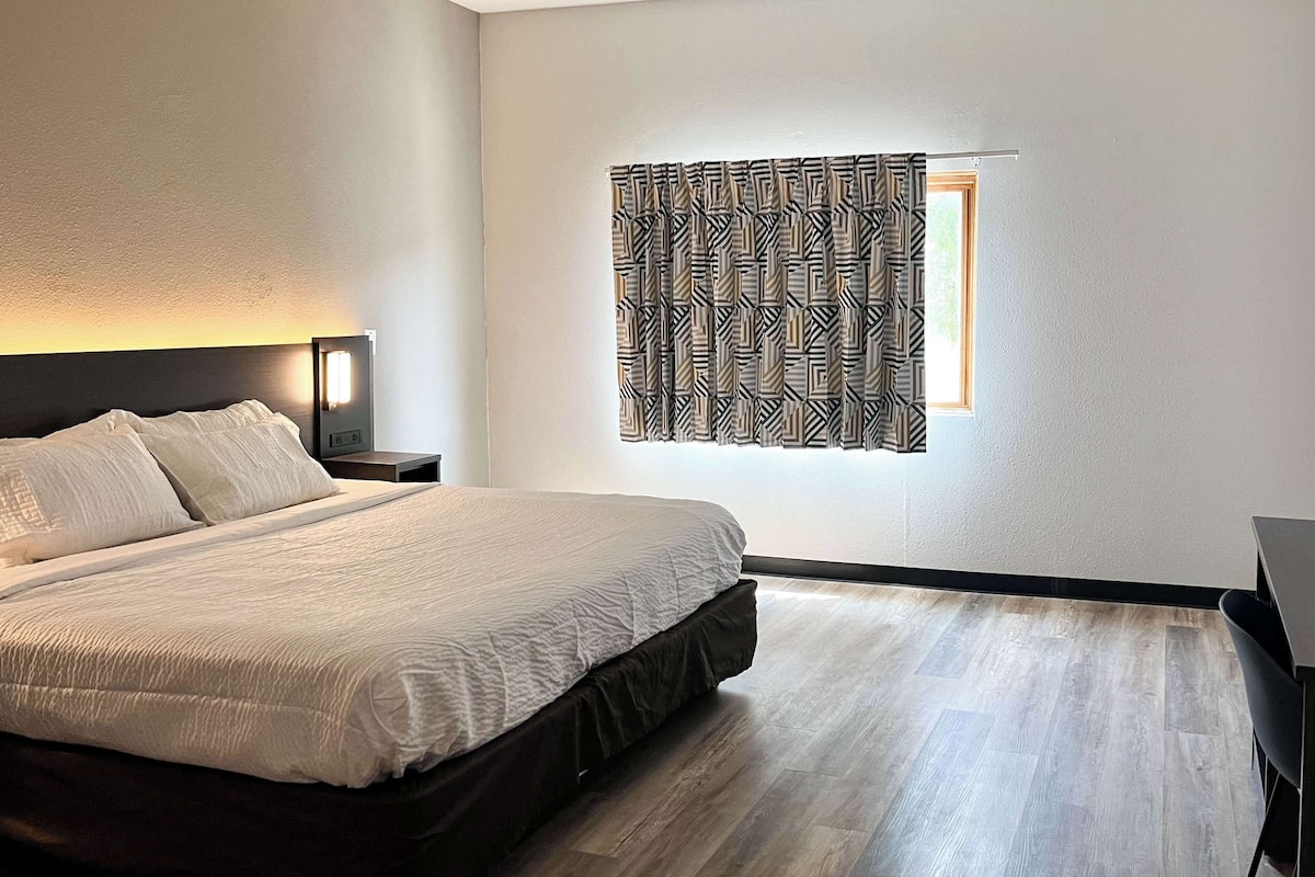 A neatly made bed with a headboard, a bedside table with a lamp, and a window with patterned curtains.