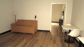 A room with a brown sofa, a white table, and a chair. There is a lamp on the left and a door leading to another room.