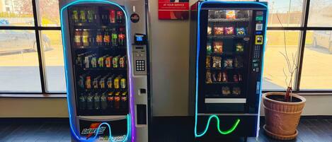 Two vending machines with a variety of snacks and drinks, placed in a room with large windows.