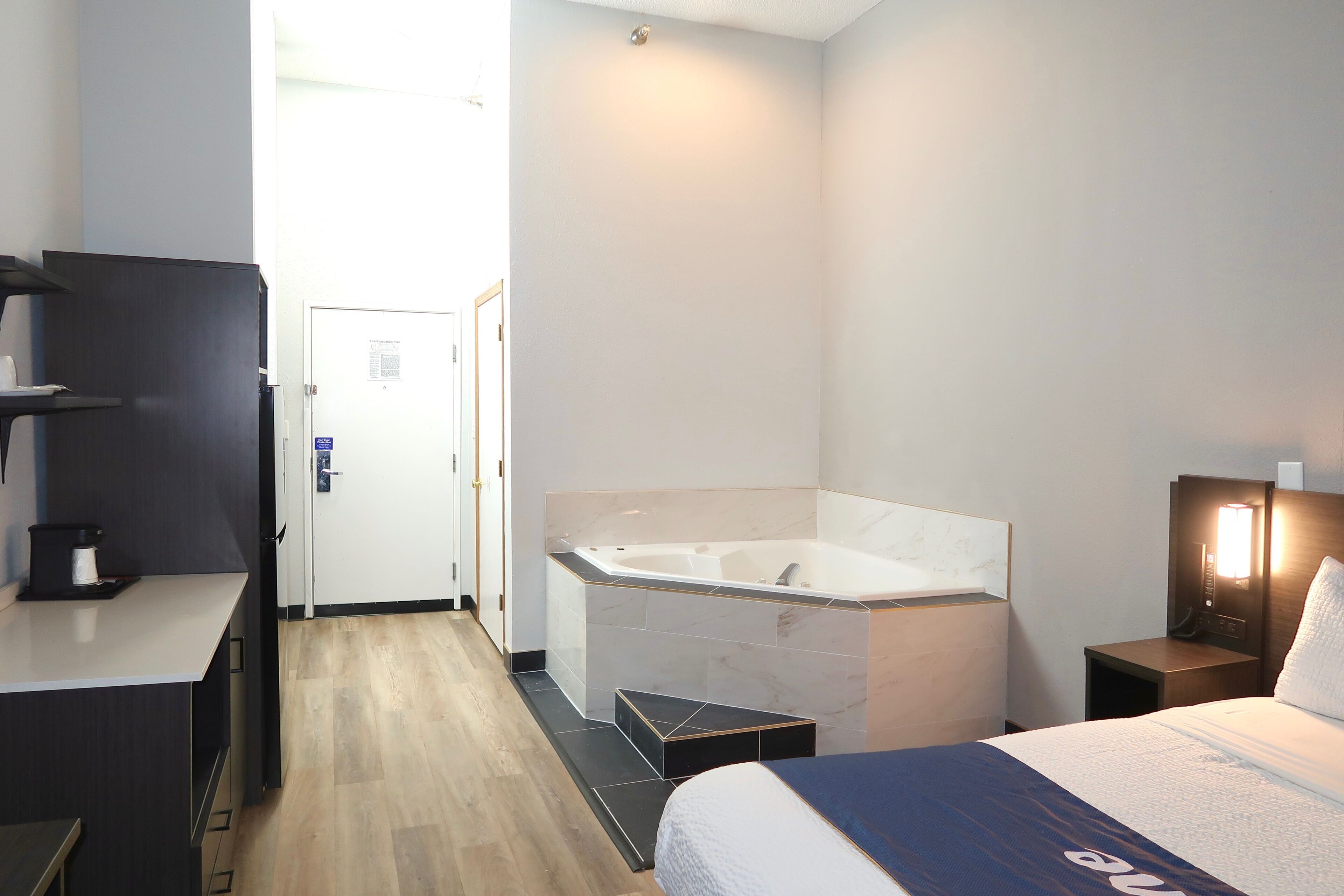 A hotel room with a large bathtub, a desk with a coffee maker, a bed with a blue and white duvet, and a nightstand with a lamp.