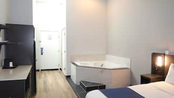 A hotel room with a large bathtub, a desk with a coffee maker, a bed with a blue and white duvet, and a nightstand with a lamp.