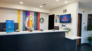 Uma receção de hotel com um grande número '6', um letreiro de 'Bem-vindo', um aviso de 'Por favor, toque no sino para assistência' e um letreiro de 'estúdio motel 6'.
