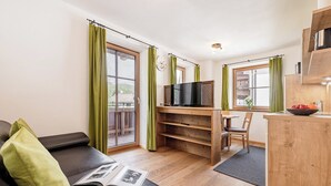 A modern living room with a sofa, a coffee table with a magazine, a TV on a wooden stand, and a dining area with a chair and table. - MYALPS Mühlhof (Oetz)