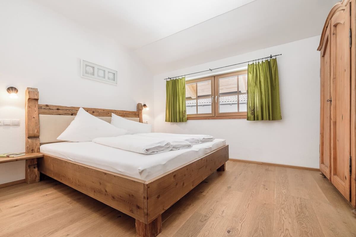 A wooden bed in a room with a window, green curtains, and a wooden wardrobe.