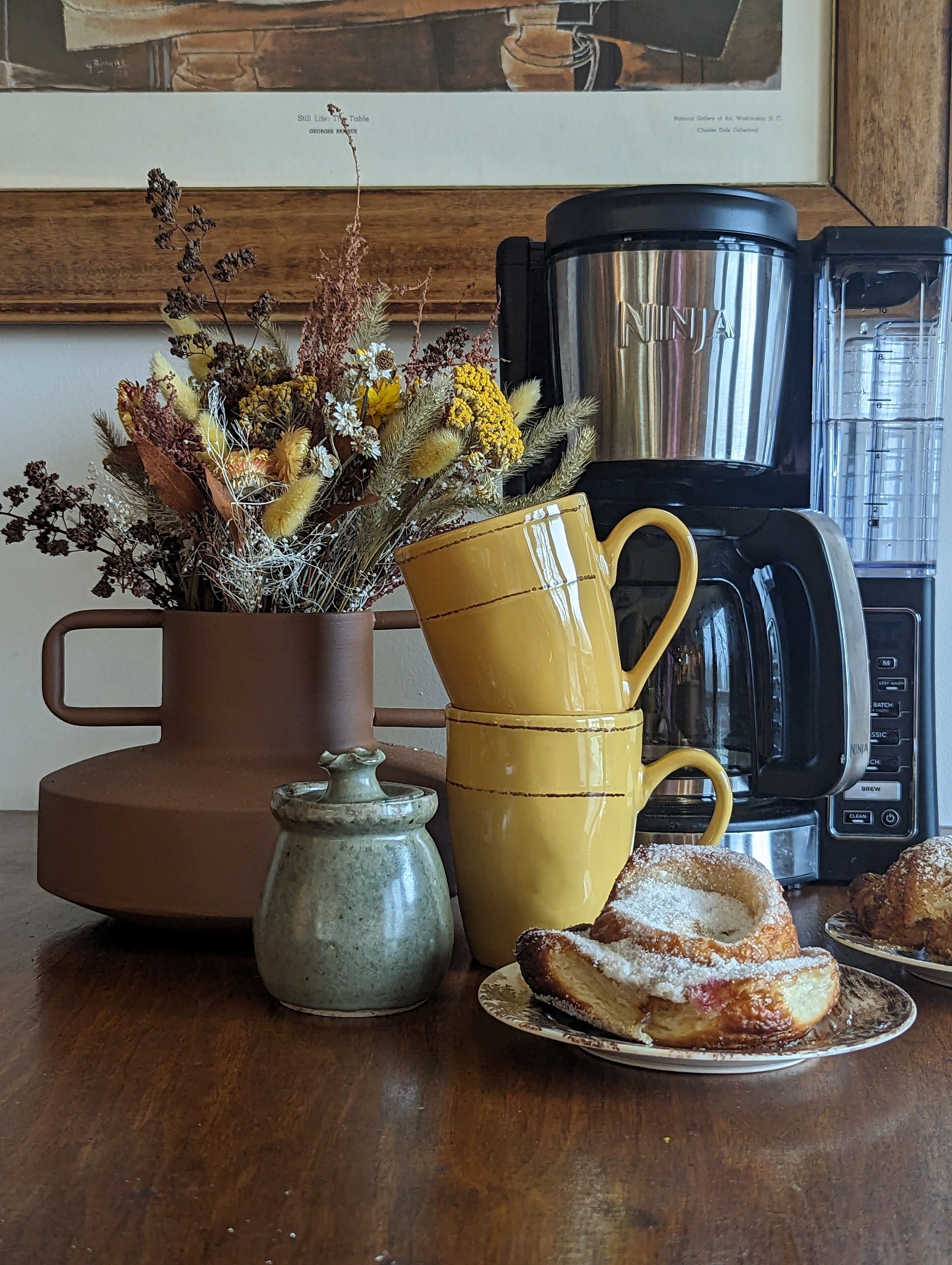 A kitchen counter with a coffee maker, a yellow mug, a ceramic pot, a vase with dried flowers, and pastries.