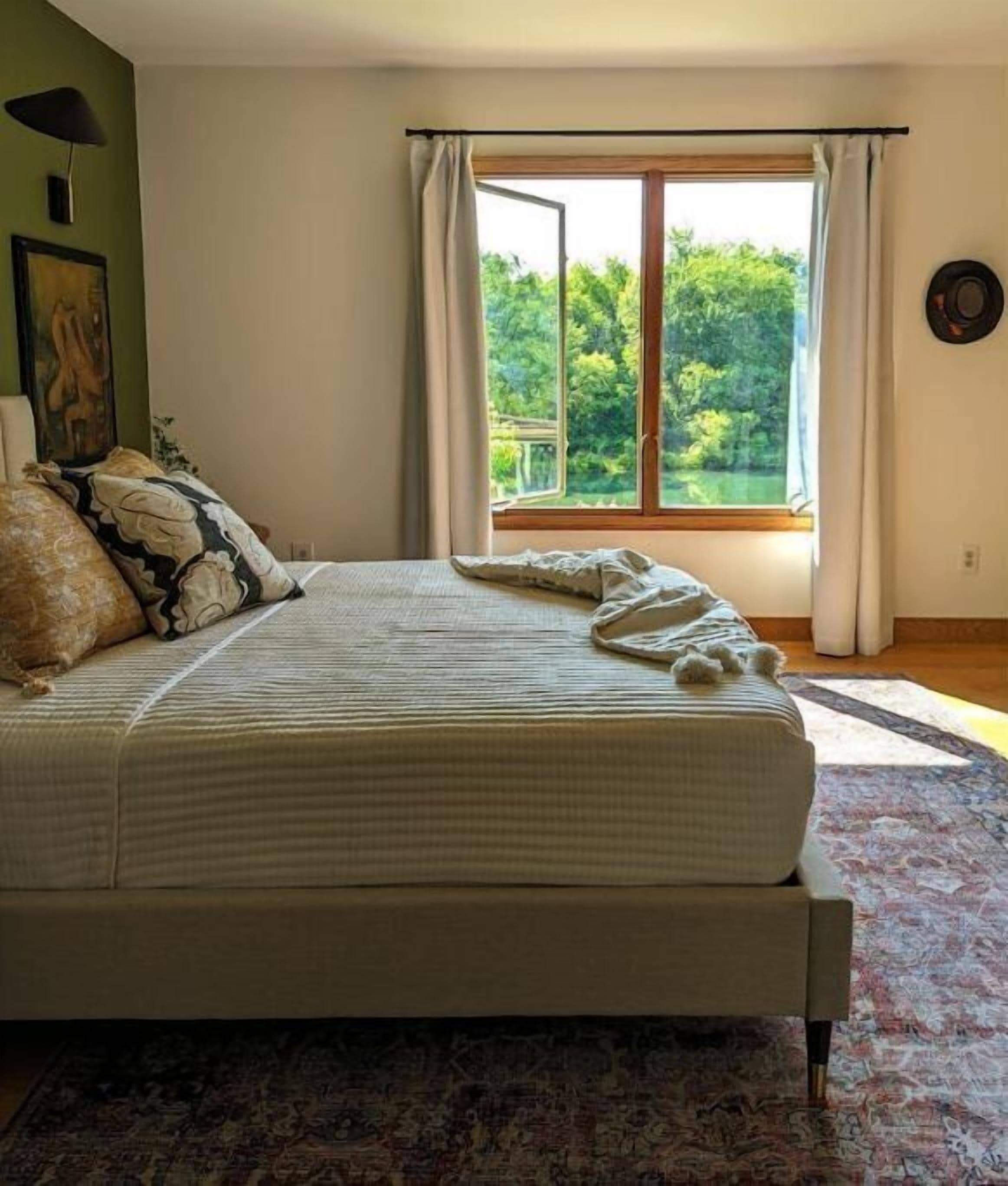 Un dormitorio con una cama grande, vistas a zonas verdes a través de una ventana, un tapete estampado y una lámpara fijada en la pared.