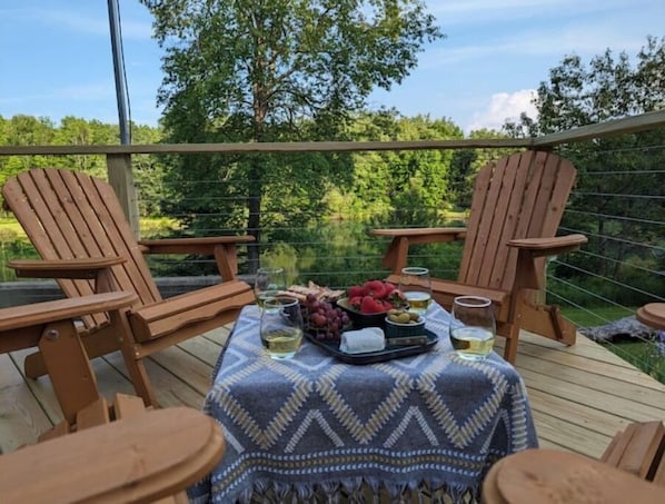 A wooden deck with two Adirondack chairs and a table set with fruits, glasses, and bottles. - Secluded Lakefront Retreat with Art Studio and Stunning Views (Otego)