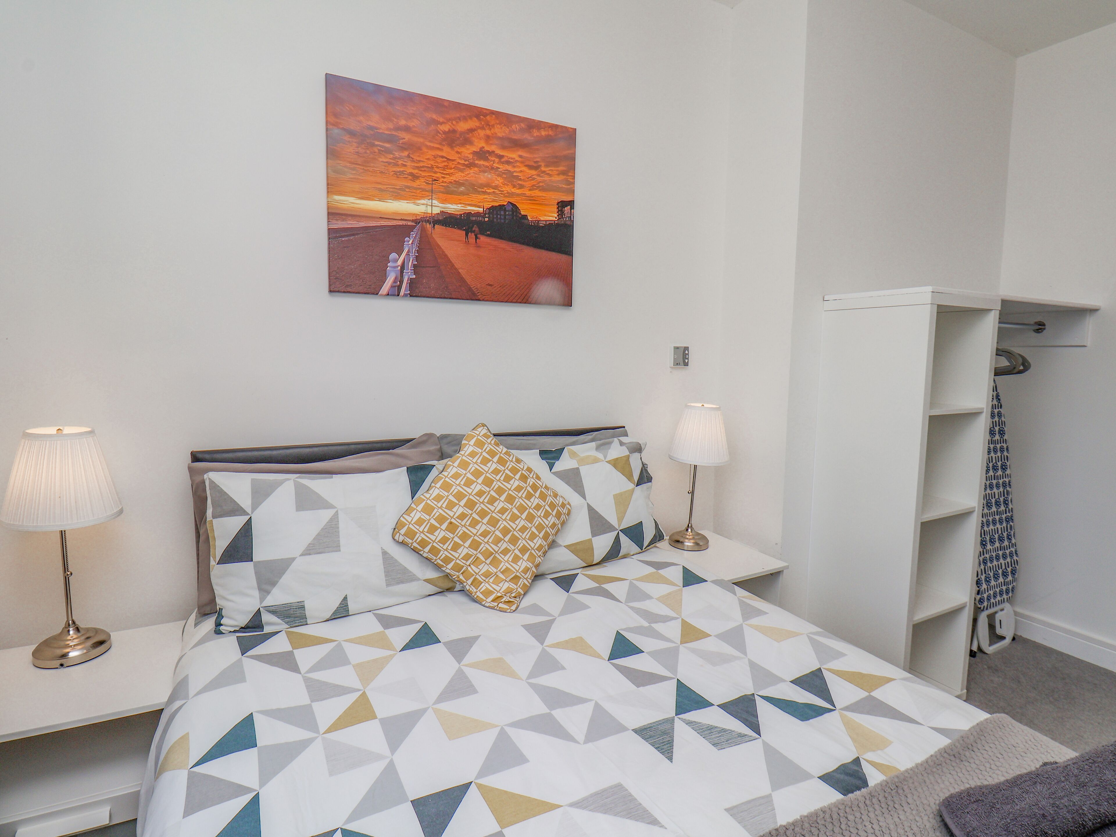 A bedroom with a bed, two bedside lamps, a geometric-patterned bedspread, a decorative pillow, a wall-mounted painting of a sunset, and a white wardrobe.