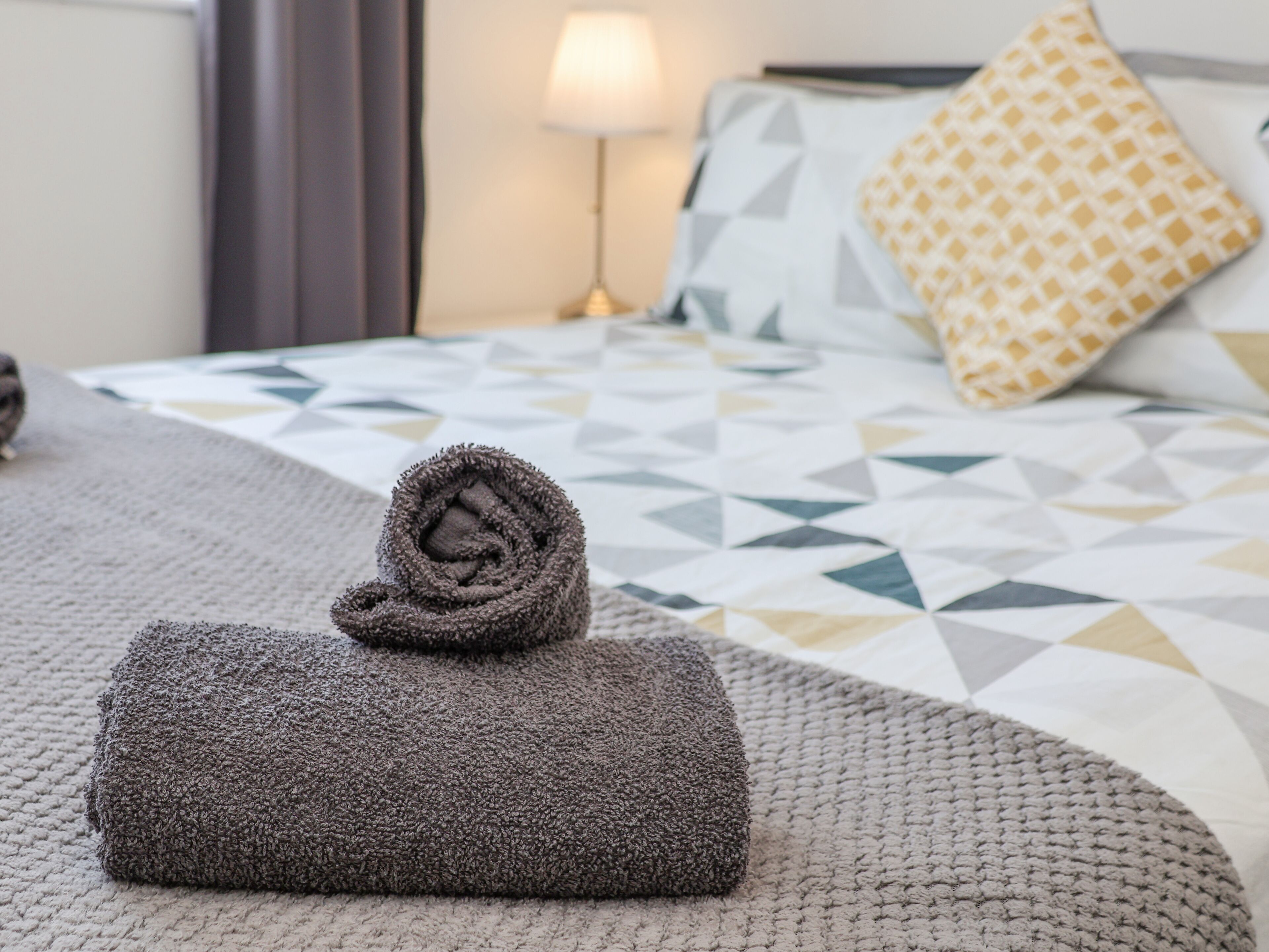 Ein ordentlich bezogenes Bett mit einem geometrisch gemusterten Überwurf, ein zusammengerolltes Handtuch auf dem Bett und eine Lampe auf dem Nachttisch.