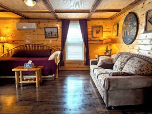 Ein Zimmer in einer Holzhütte mit einem Bett, einem Schlafsofa, einem Tisch und einem Fenster mit Vorhängen.