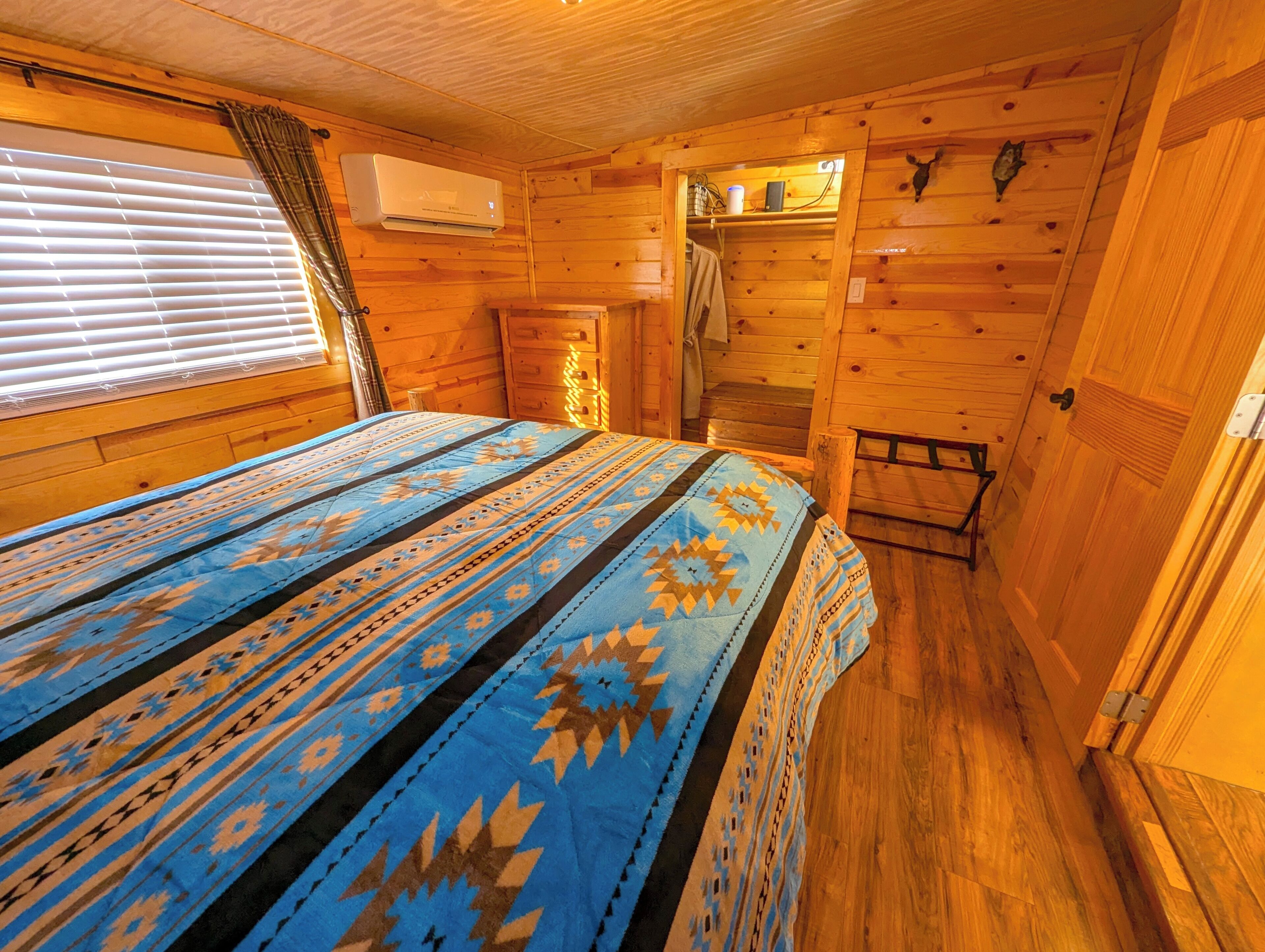 Une chambre dans un chalet en bois, comprenant un lit, une fenêtre avec stores, un appareil de climatisation et un escalier menant à un étage supérieur.
