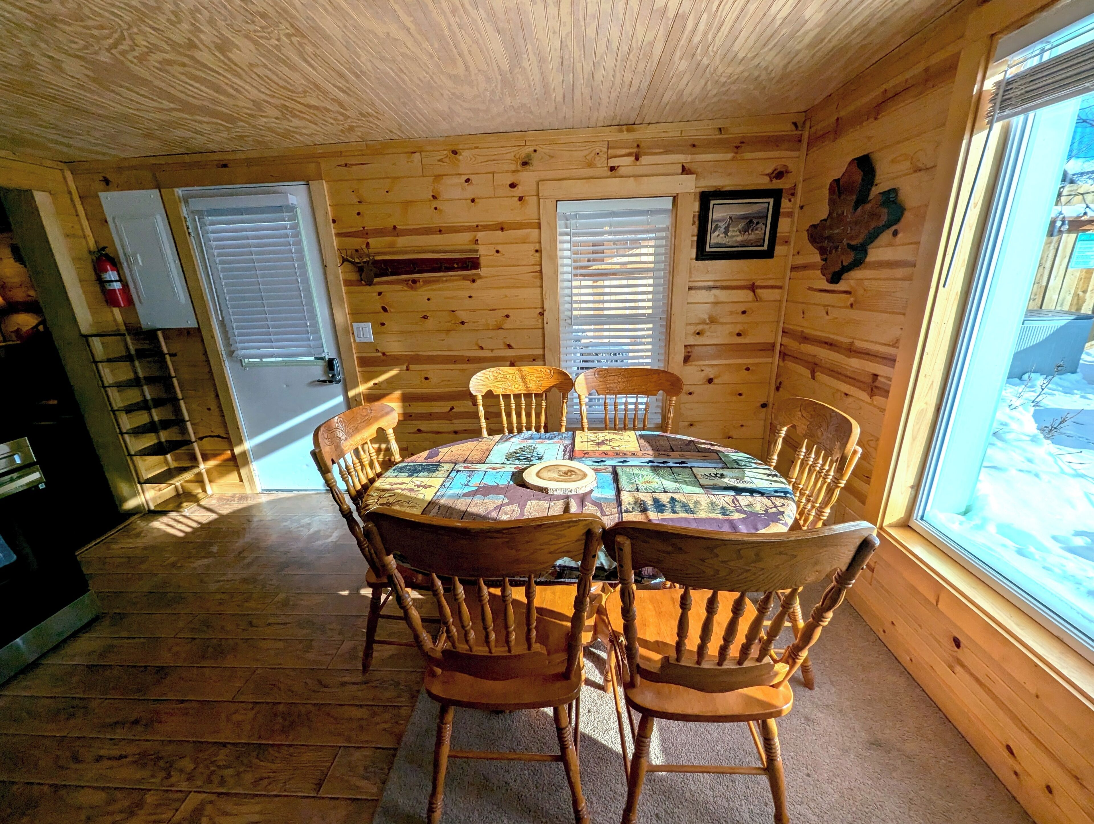 A wooden cabin interior with a round dining table, six wooden chairs, and a window with blinds.