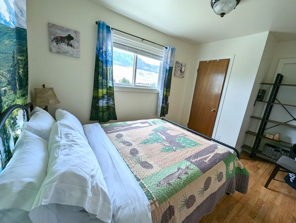 A bedroom with a bed, a window with curtains, a door, a lamp, and a shelf with books.