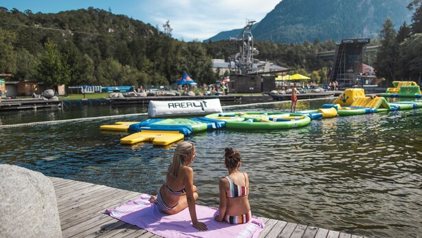 A wooden dock with a pink towel, a water body with inflatable rafts, and a mountainous backdrop.