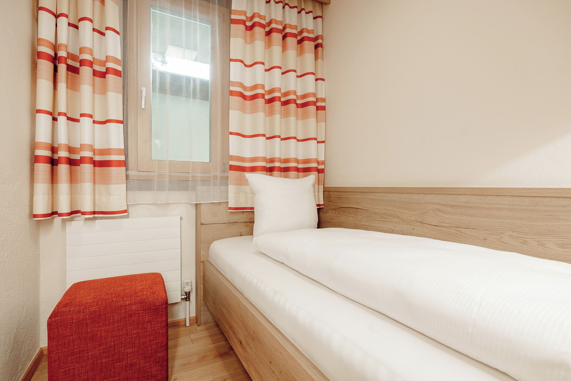 A bedroom with a bed, a window with curtains, a radiator, and a red bench.