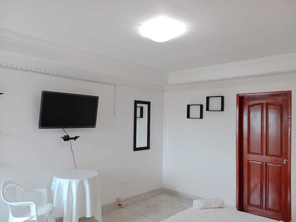 A room with a flat-screen TV mounted on the wall, a small round table with a chair, a wooden door, and a mirror.