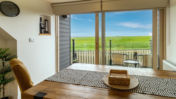 A dining area with a wooden table, a patterned rug, and a view of a green field through large sliding glass doors. - Picturesque cottage with views (Warmfield)