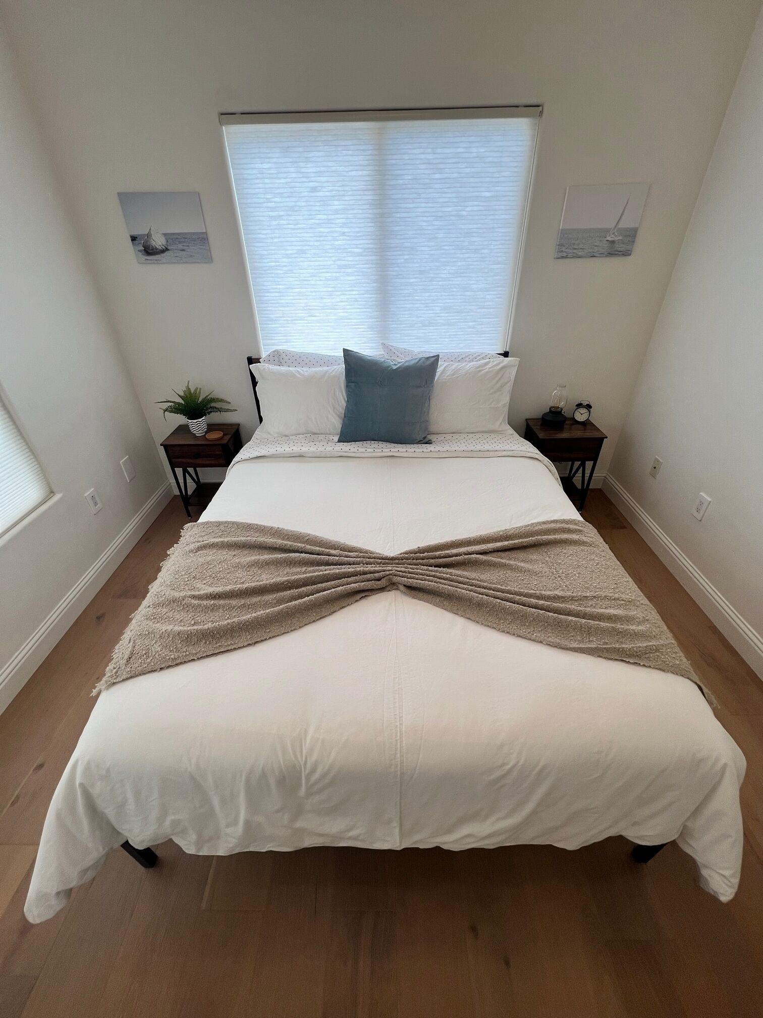 A large bed with a white and beige bedspread, two pillows, a small table with a plant, and a nightstand with a clock.