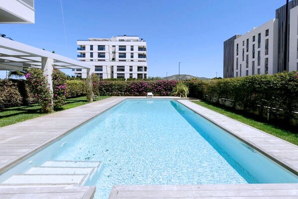 A rectangular swimming pool with clear blue water, surrounded by a wooden deck and bordered by hedges. In the background, there are modern buildings with balconies.