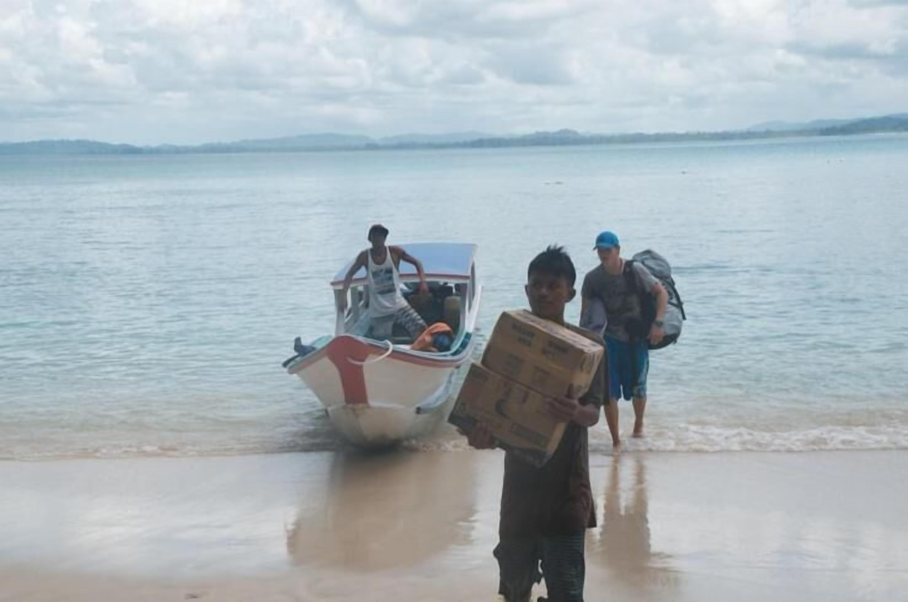 A boat on a calm body of water with a person standing on the shore holding a box.