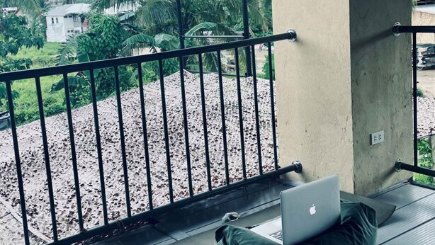 Un balcon avec un ordinateur portable, une bouteille d’eau et une tasse, donnant sur un espace recouvert de gravier, avec des palmiers et des maisons en arrière-plan.