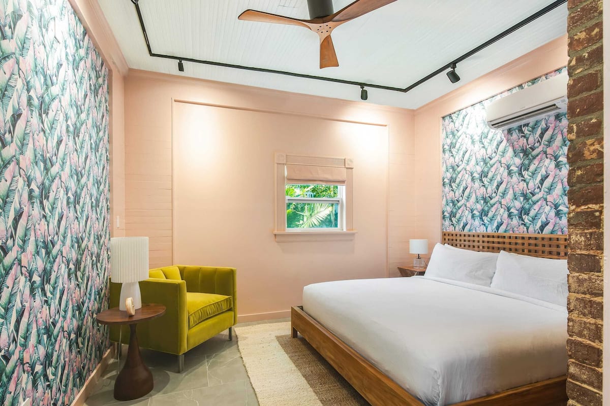 A bedroom with a bed, a green armchair, a small table, a window, and a ceiling fan.