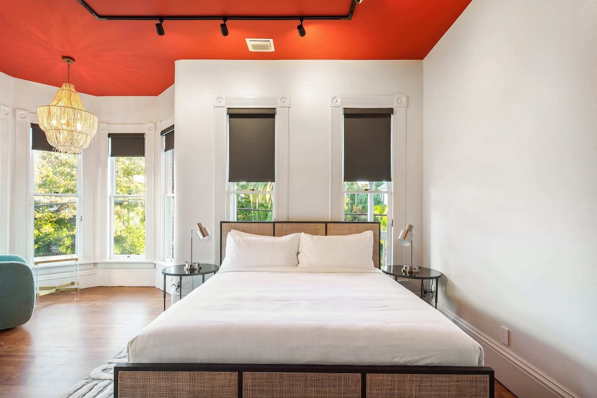 A bedroom with a large bed, two nightstands, a hanging light fixture, and windows with blinds.