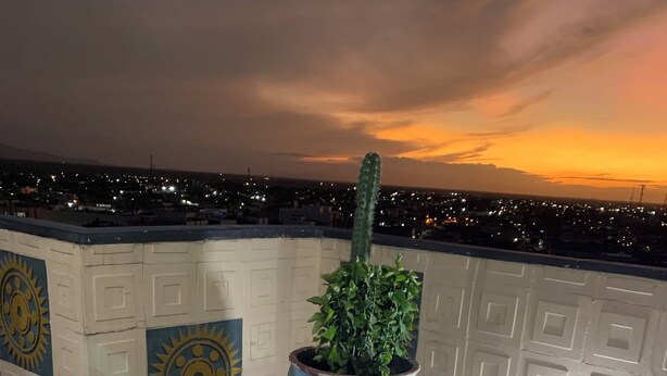 Une terrasse avec une table, des chaises, un pot de fleurs et une vue sur la ville au coucher du soleil.