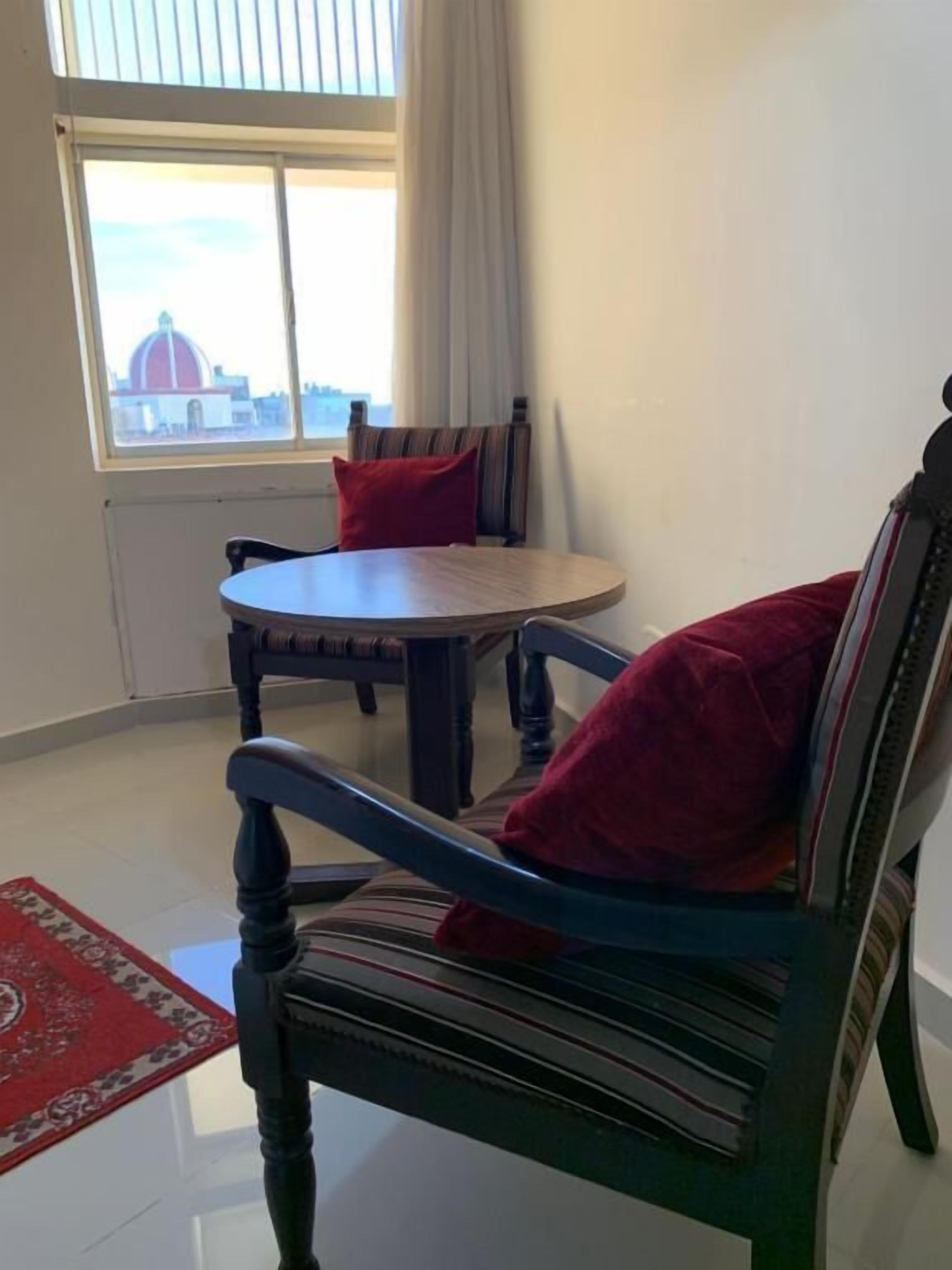 Una stanza con un tavolo rotondo e due sedie, un cuscino rosso su una sedia, una finestra con vista su un edificio e un tappeto rosso a motivi sul pavimento.