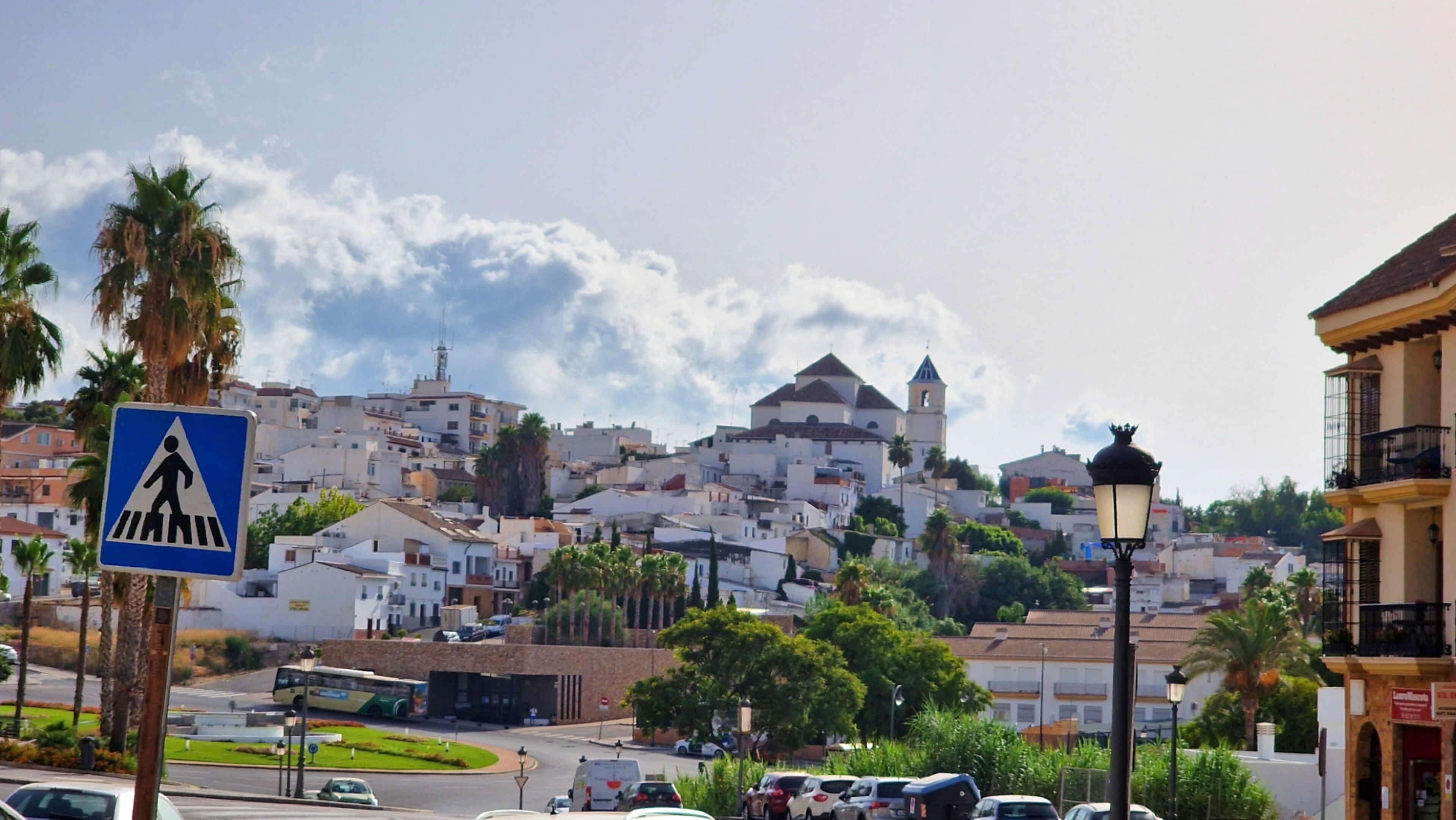 A town with white buildings, a pedestrian crossing sign, and a street with cars and a bus.