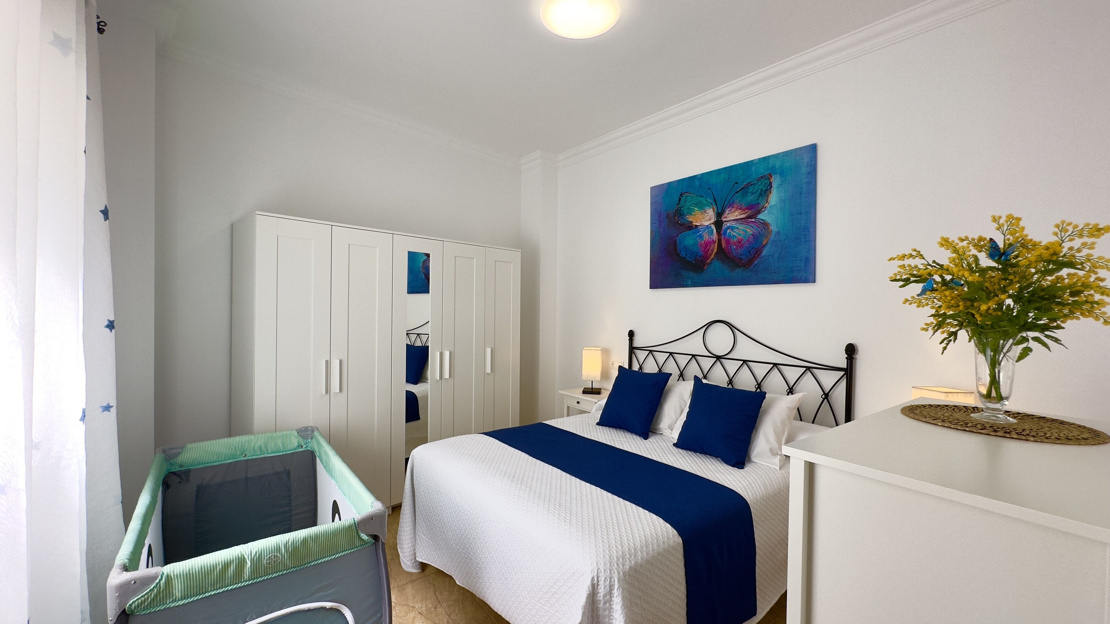 A bedroom with a bed, a wardrobe, a crib, a painting, and a vase with flowers.