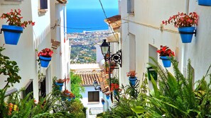 A narrow street with white buildings, blue flower pots, and a view of a city and sea in the distance. - Apartamento Céntrico La Abuela Cati (Alhaurín el Grande)