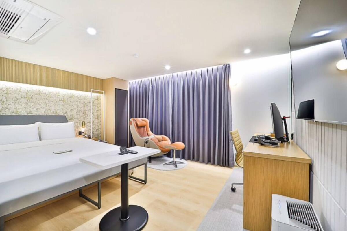 A modern hotel room with a large bed, a desk with a computer, a chair, and a large window with curtains.