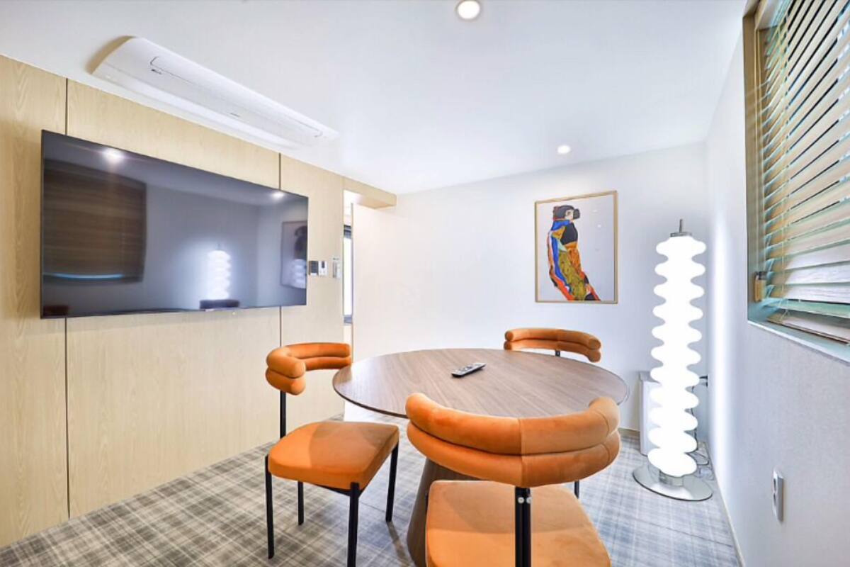 A modern dining area with a round table, orange upholstered chairs, a flat-screen TV mounted on the wall, and a painting on the wall.