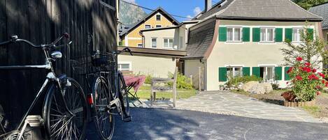 Una bicicleta estacionada frente a un edificio de madera, un camino pavimentado que conduce a una casa con contraventanas verdes, y una montaña al fondo.