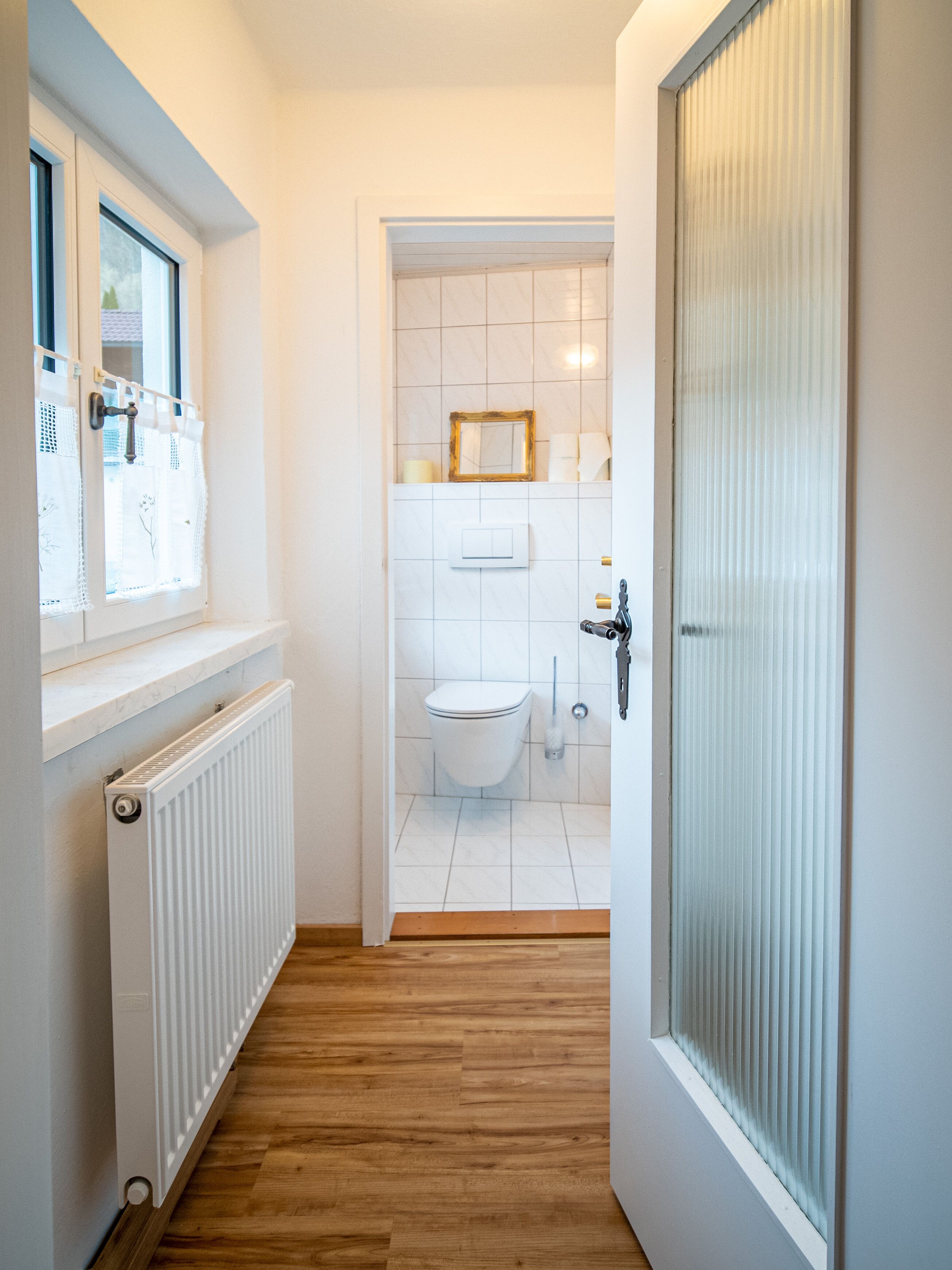A narrow hallway leading to a bathroom with a toilet, a radiator, and a window with a curtain.