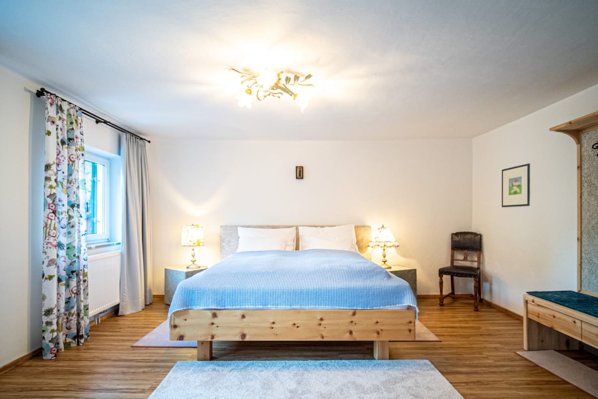 A bedroom with a wooden bed, two bedside lamps, a window with floral curtains, a wooden chair, and a framed picture on the wall.