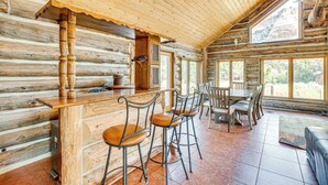 A log cabin interior with a bar area, wooden chairs, and large windows offering views of the outdoors.
