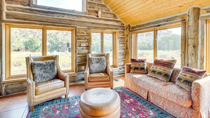 A log cabin interior with large windows, a leather armchair, a sofa with patterned cushions, and a round ottoman.
