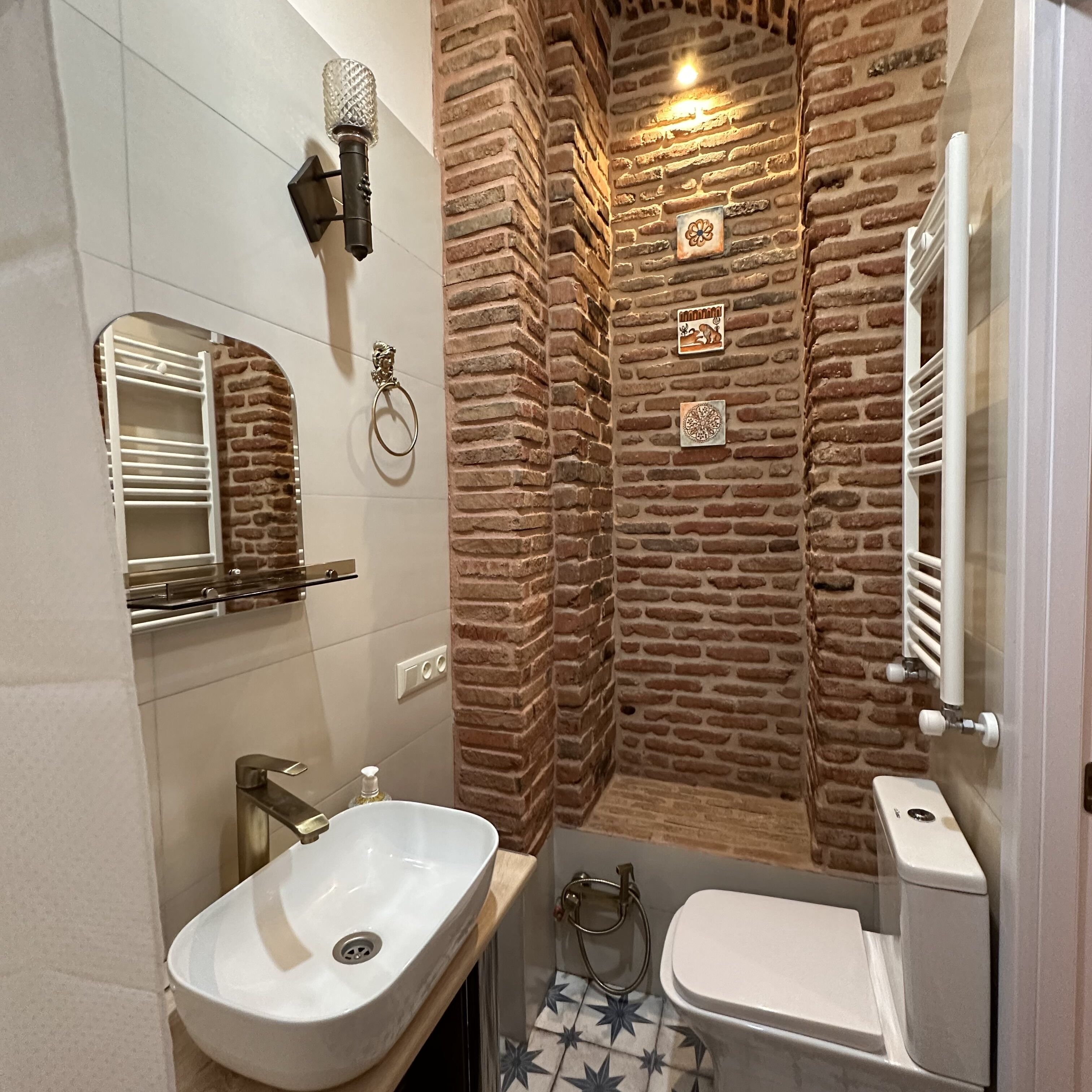 Un baño con un lavamanos blanco, un inodoro y una zona de ducha. Hay un espejo sobre el lavamanos y un perchero. Las paredes están decoradas con un diseño de ladrillo.